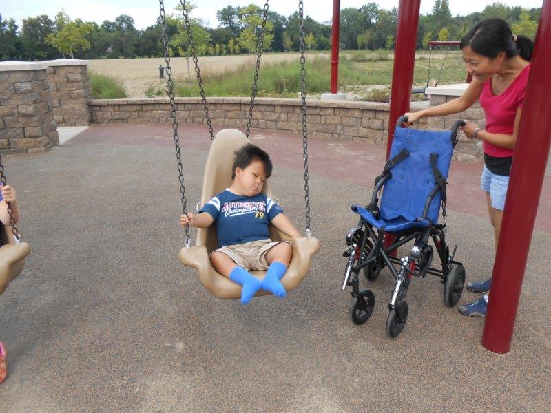 A mom sets a wheelchair adjacent to the swing in preparation to assist her young son in transfer from the swing to his wheelchair.