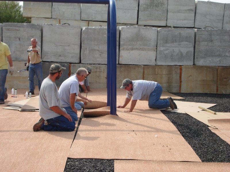 A park maintenance crew of four men are shown on their knees seaming together the top mat of a hybrid surface system as it is installed.