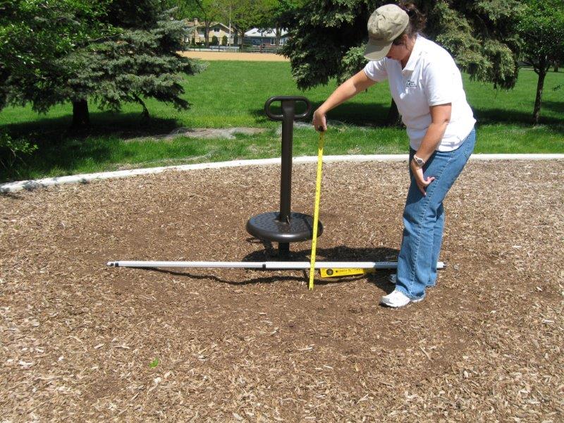 A researcher measures the depth of a kick-out area at a ground level play component with a two meter straight edge laid horizontal over the surface.