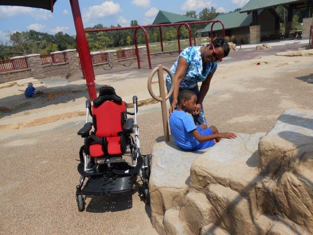 A mom assists her young son to move from his wheelchair to seated on the climbing rock-like transfer system.