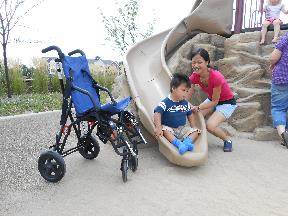 A little boy comes down the slide as he is assisted by his mom.  A wheelchair is seated next to the bottom of the slide.