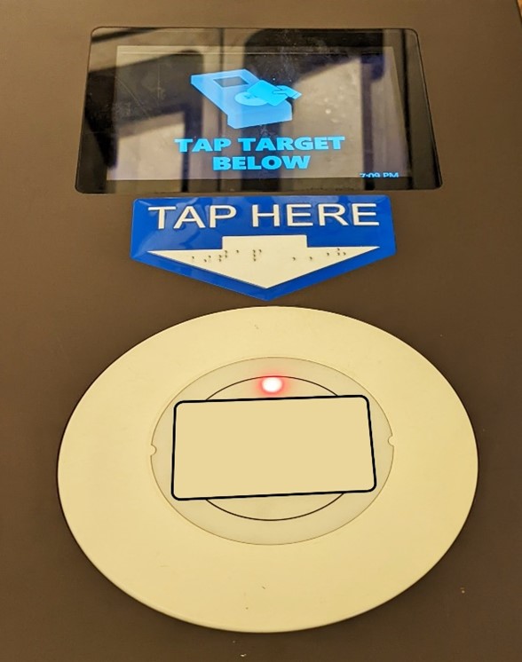 Raised contactless payment card reader with red indicator lights. Above is a tactile sticker that says tap here with an arrow pointing down and rail on the arrow. At the top is an LCD screen that says tap target below.